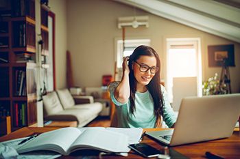 A student smiles as she works at a laptop in a cozy bedroom or loft space.